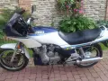 Klasyczny motocykl Yamaha XJ 900 w kolorze białym i granatowym, z niebieskimi i żółtymi pasami, zaparkowany na brukowanym podjeździe.