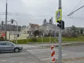 Szare auto zbliża się do przejścia dla pieszych, gdzie światła kolizyjne regulują ruch. W tle budynki i drzewa.