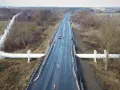 Duży biały rurociąg biegnie wzdłuż drogi krajowej 91. Widać samochody na mokrej nawierzchni i drzewa.