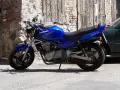 Błękitny motocykl Kawasaki ER-5 zaparkowany przy kamiennym murze i drewnianej bramie.