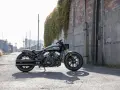 Czarny motocykl typu cruiser Indian Scout stoi na ulicy, z zielonym bluszczem zwisającym ze ściany w tle.