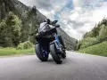 Motocyklista na Yamaha Niken pokonuje zakręt na górskiej drodze, otoczony zielonymi lasami i skalistymi szczytami.