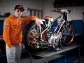 Młody żużlowiec w pomarańczowej bluzie opiera się o swój motocykl żużlowy, gotowy do kolejnego wyścigu.