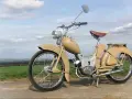 Beżowy motorower z NRD, z charakterystycznym siodełkiem i błotnikami, stoi na tle zielonych pól i błękitnego nieba.