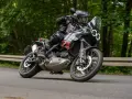 Motocyklista w kasku i stroju terenowym pochyla się na zakręcie na drodze leśnej, prowadząc czarny motocykl Ducati DesertX.