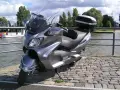 Szary Suzuki Burgman 650, gotowy na podróż w stylu V-max, stoi nad wodą.