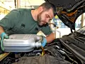 Mechanik w zielonym kombinezonie i niebieskich rękawiczkach nalewa olej do silnika. W tle widać napis "2 t".