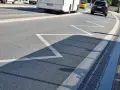 Na drodze zygzakowata linia, znak p-17, zapowiada zbliżanie się do przejścia dla pieszych. W tle autobus i samochód.