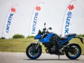 Niebieski motocykl Suzuki GSX-S z niebieskimi felgami stoi na asfalcie. W tle powiewają białe flagi z logo Suzuki.