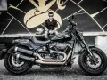 Mężczyzna w czarnej koszuli i jeansach siedzi obok lśniącego, czarnego motocykla Harley-Davidson Fat Bob.
