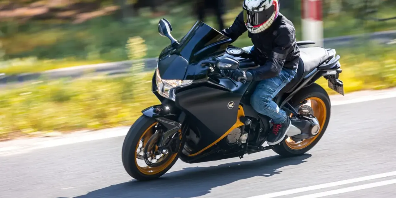 Motocyklista w kasku z uśmiechniętą twarzą pędzi na czarnym motocyklu Honda VFR 1200 po drodze.