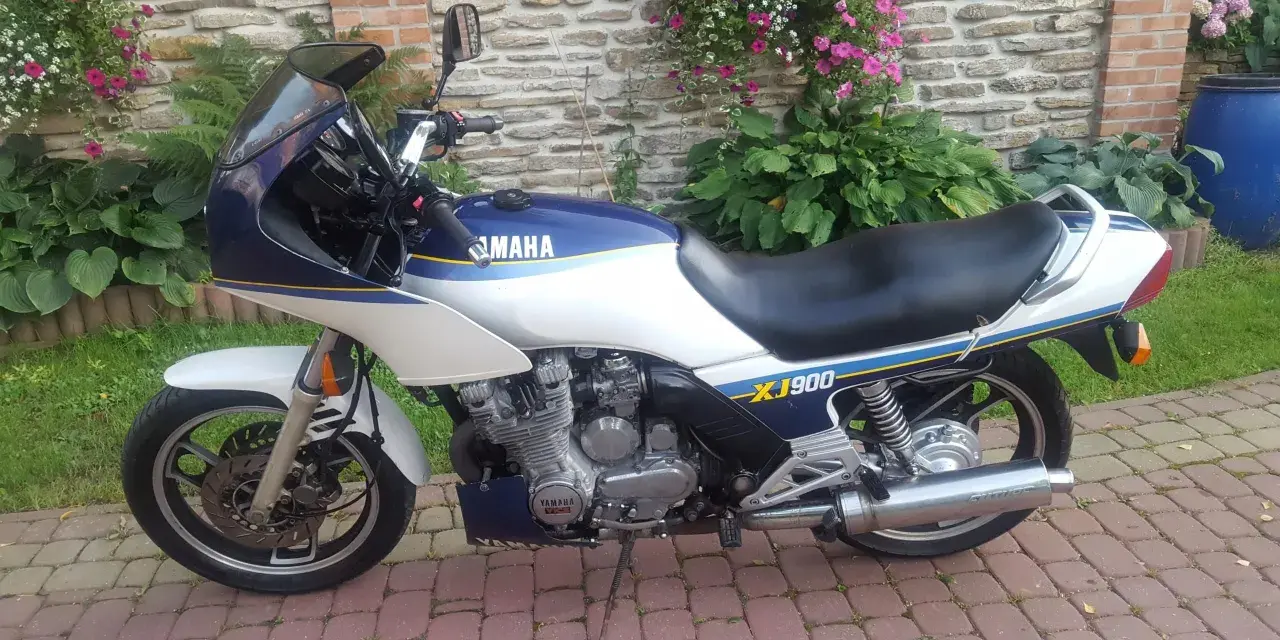 Klasyczny motocykl Yamaha XJ 900 w kolorze białym i granatowym, z niebieskimi i żółtymi pasami, zaparkowany na brukowanym podjeździe.