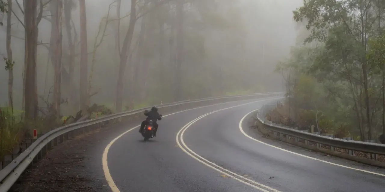Motocyklista pokonuje zakręt na mokrej drodze, gdzie grozi mu aquaplaning. Mgła otula las, tworząc tajemniczą atmosferę.
