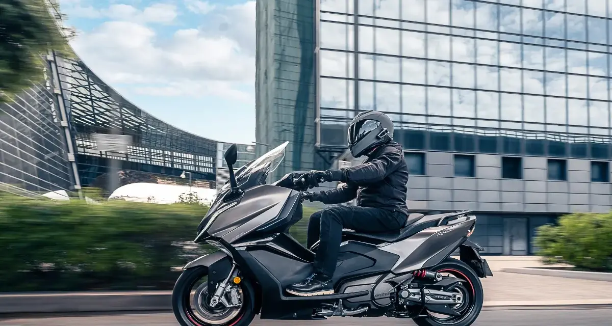 Mężczyzna w kasku jedzie na skuterze Kymco AK 550 obok nowoczesnego budynku.