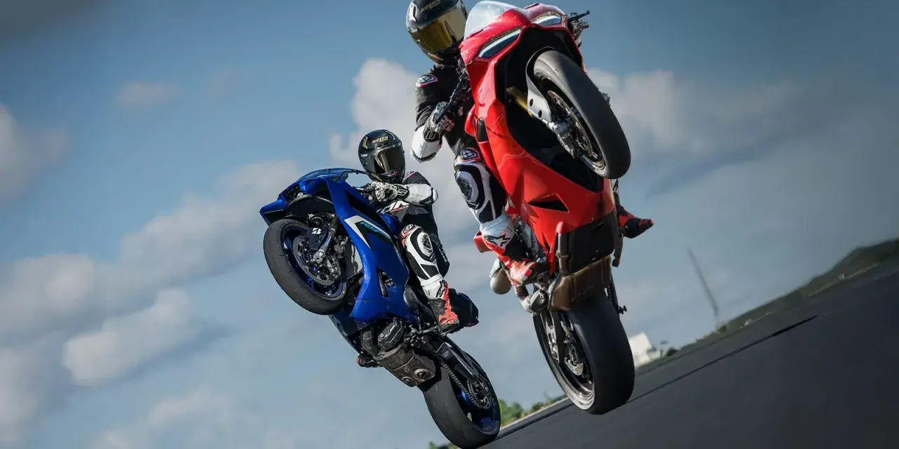 Dwóch motocyklistów na torze wyścigowym wykonuje wheelie. Jeden na niebieskim motocyklu, drugi na czerwonym Ducati Panigale V2.