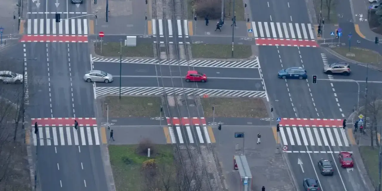 Skrzyżowanie z tramwajami i pasami. Czerwony samochód wykonuje wyprzedzanie na pasach, ryzykując kolizję z pieszymi.