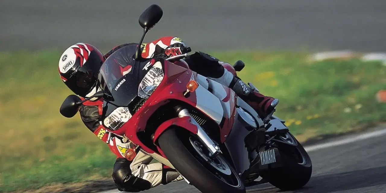 Motocyklista na torze pochyla się na swoim czerwono-białym motocyklu Yamaha R6 2001, w pełni ubrany w kombinezon i kask.