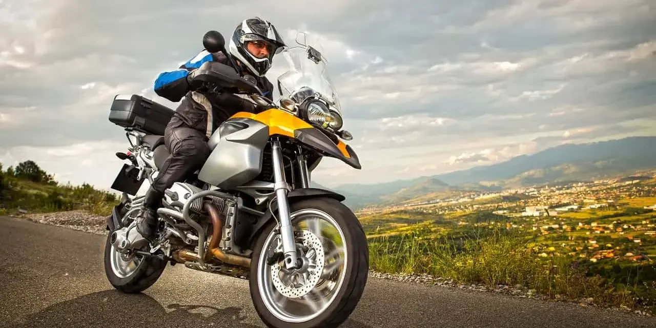 Motocyklista w kasku jedzie na motocyklu turystycznym. W tle rozciąga się malowniczy krajobraz z miastem. Zastanawiasz się nad koszt przerejestrowania motocykla?