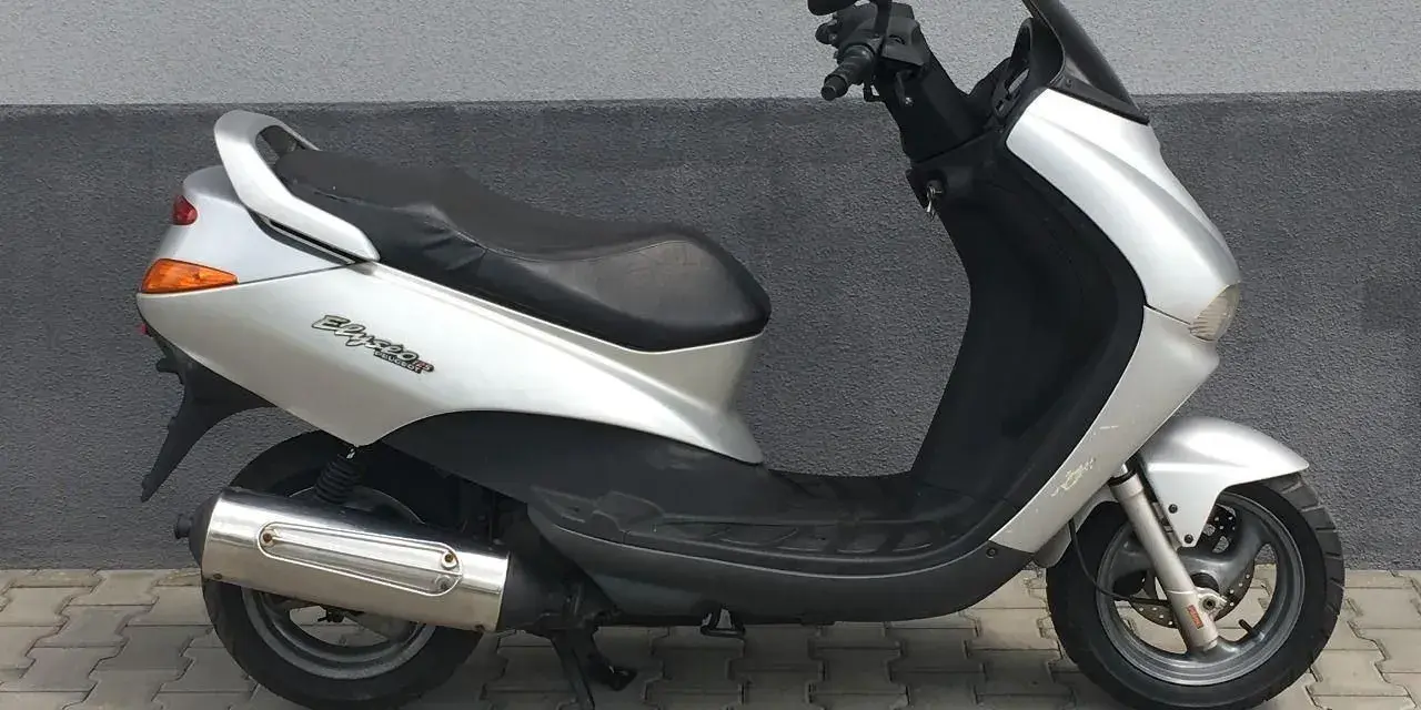 Srebrny skuter Peugeot Elyseo 125 z czarnym siedzeniem i przyciemnianą szybą, zaparkowany na chodniku.