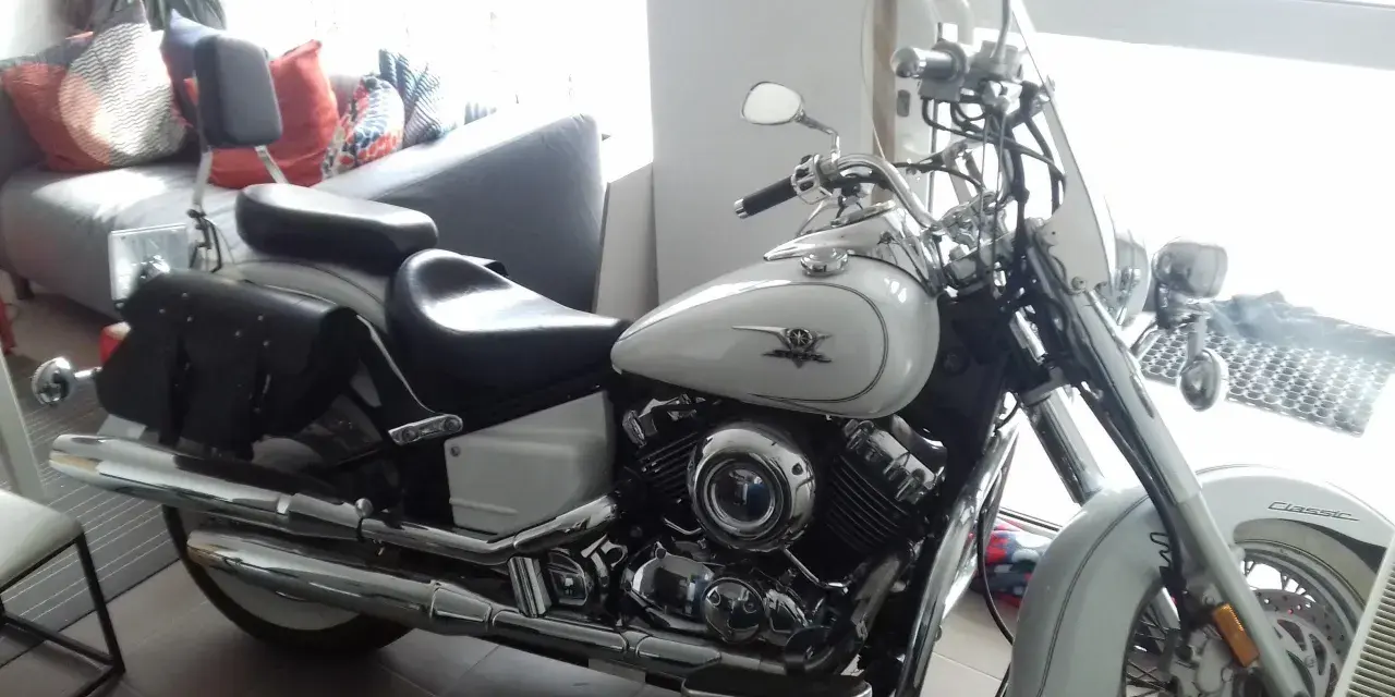 Biały motocykl Yamaha Drag Star 650, z sakwami i szybą. Wady i zalety tego klasyka są dyskutowane w tle, gdzie widać kanapę.