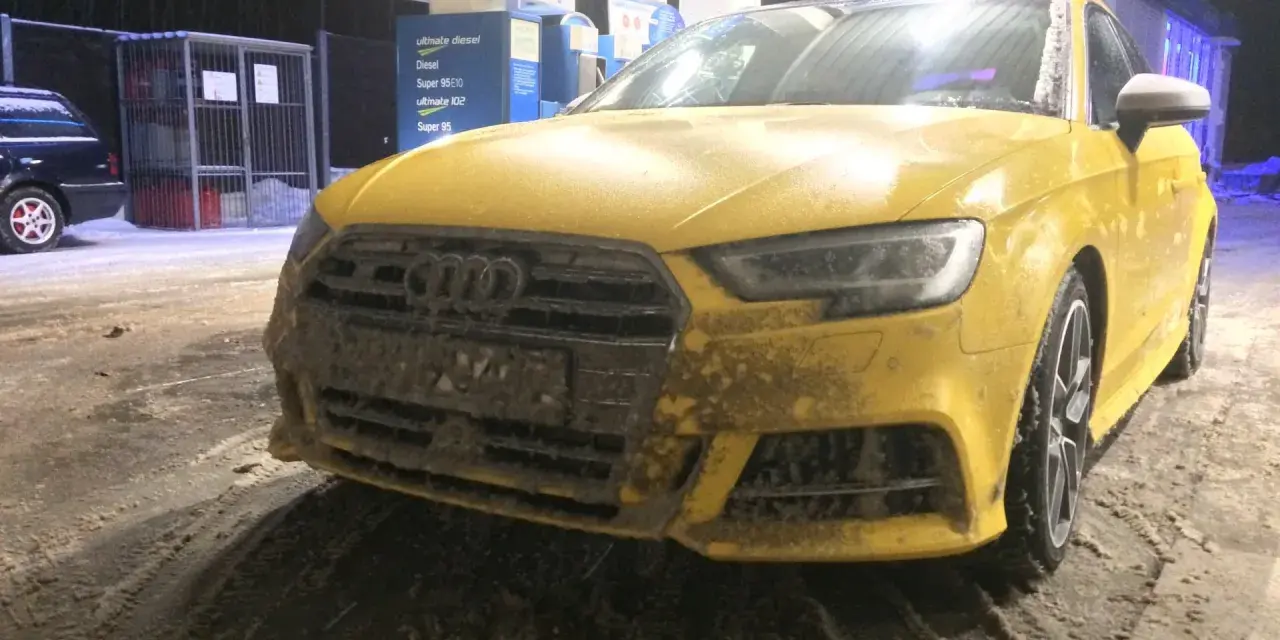 Żółte Audi na stacji benzynowej, pokryte błotem i śniegiem. Nieczytelna tablica rejestracyjna utrudnia identyfikację.