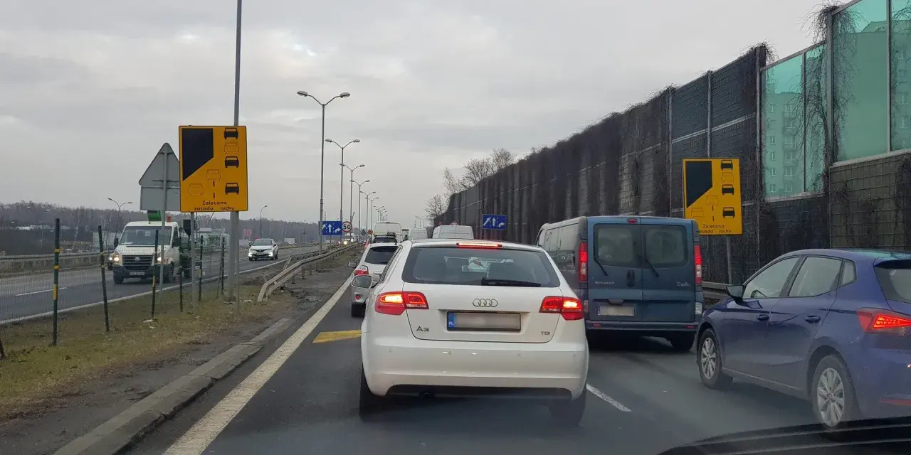 Korek na drodze, znaki informują o zalecanej zasadzie ruchu "na suwak". Unikaj mandatu, stosuj się do przepisów.