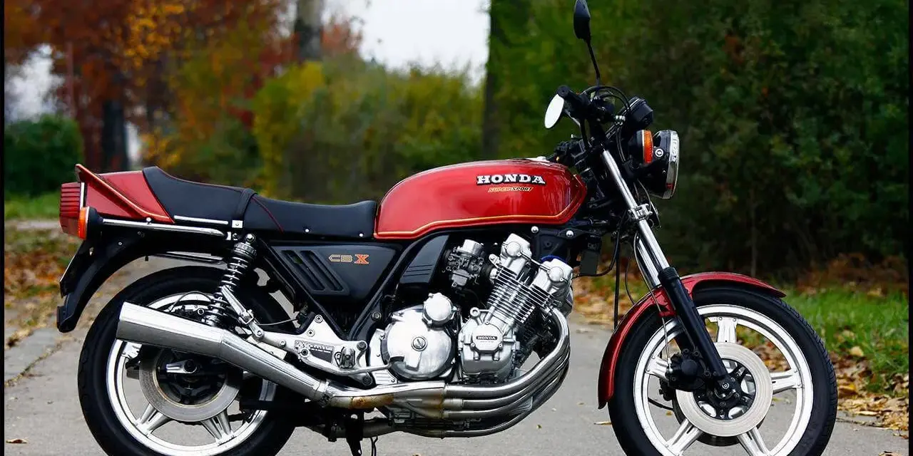 Klasyczny motocykl Honda CBX 1000 w kolorze czerwonym, z widocznym potężnym silnikiem i chromowanymi elementami, zaparkowany na tle jesiennej zieleni.