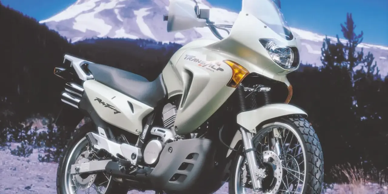 Srebrny motocykl Honda Transalp 650 gotowy na przygodę na tle ośnieżonej góry i lasu.
