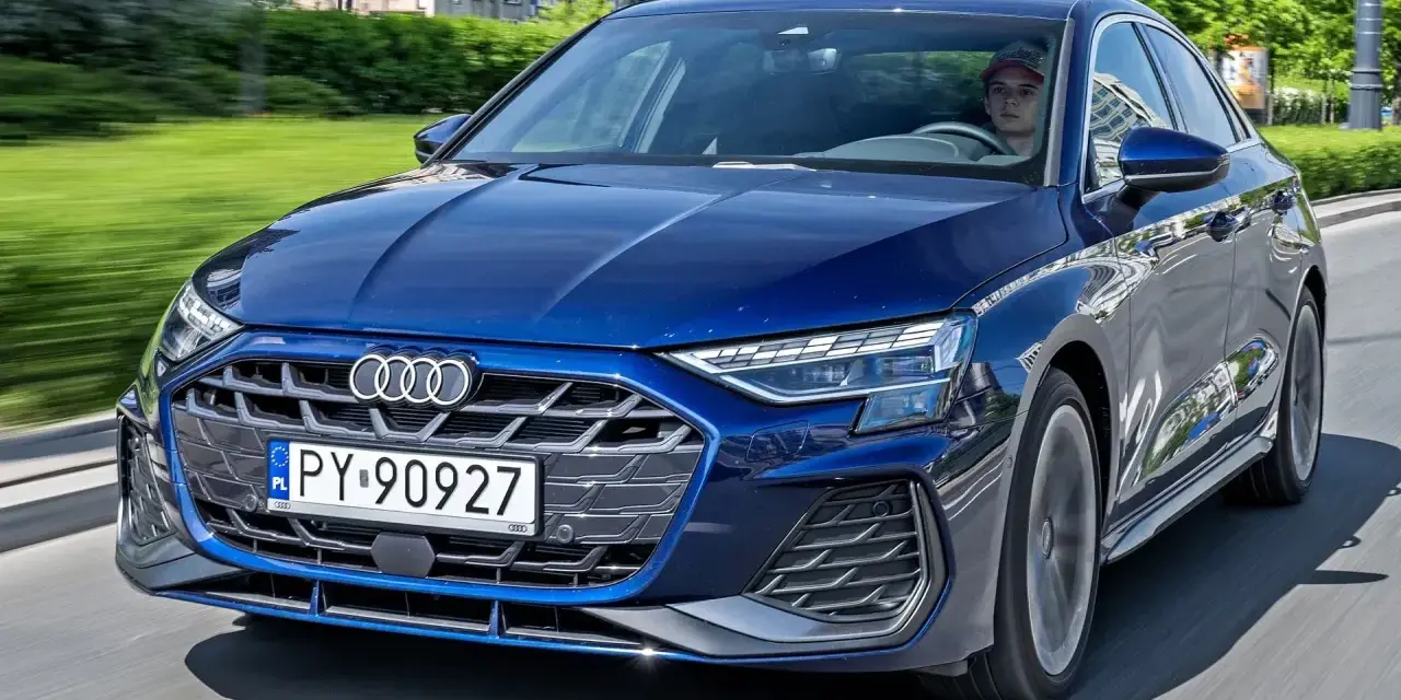 Młody kierowca w czapce za kierownicą granatowego Audi A3, jadącego po miejskiej drodze.