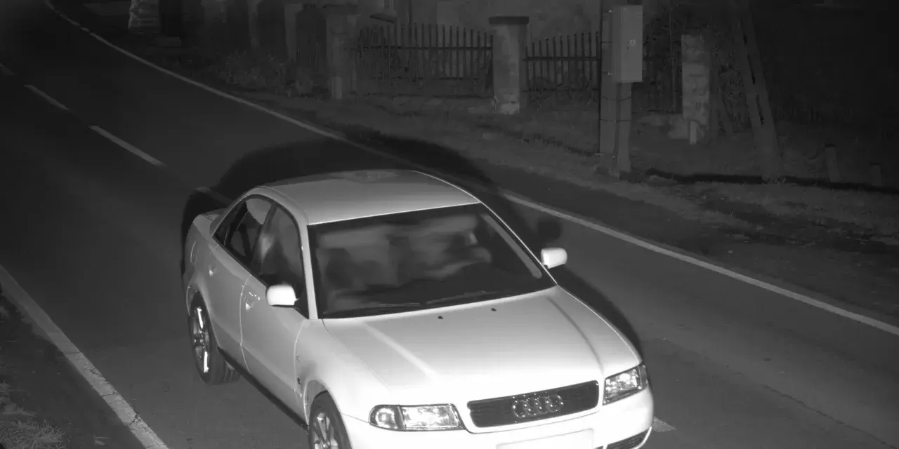 Biały sedan Audi przejeżdża przez nocne zdjęcie z fotoradaru.
