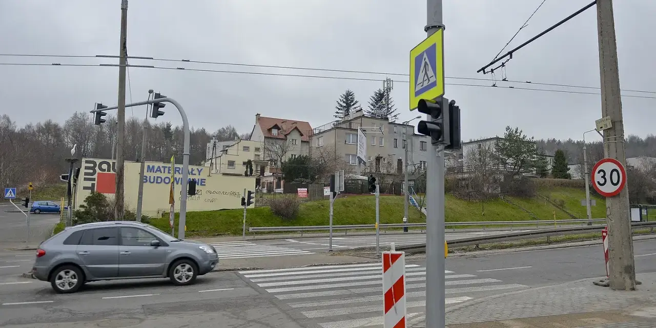 Szare auto zbliża się do przejścia dla pieszych, gdzie światła kolizyjne regulują ruch. W tle budynki i drzewa.