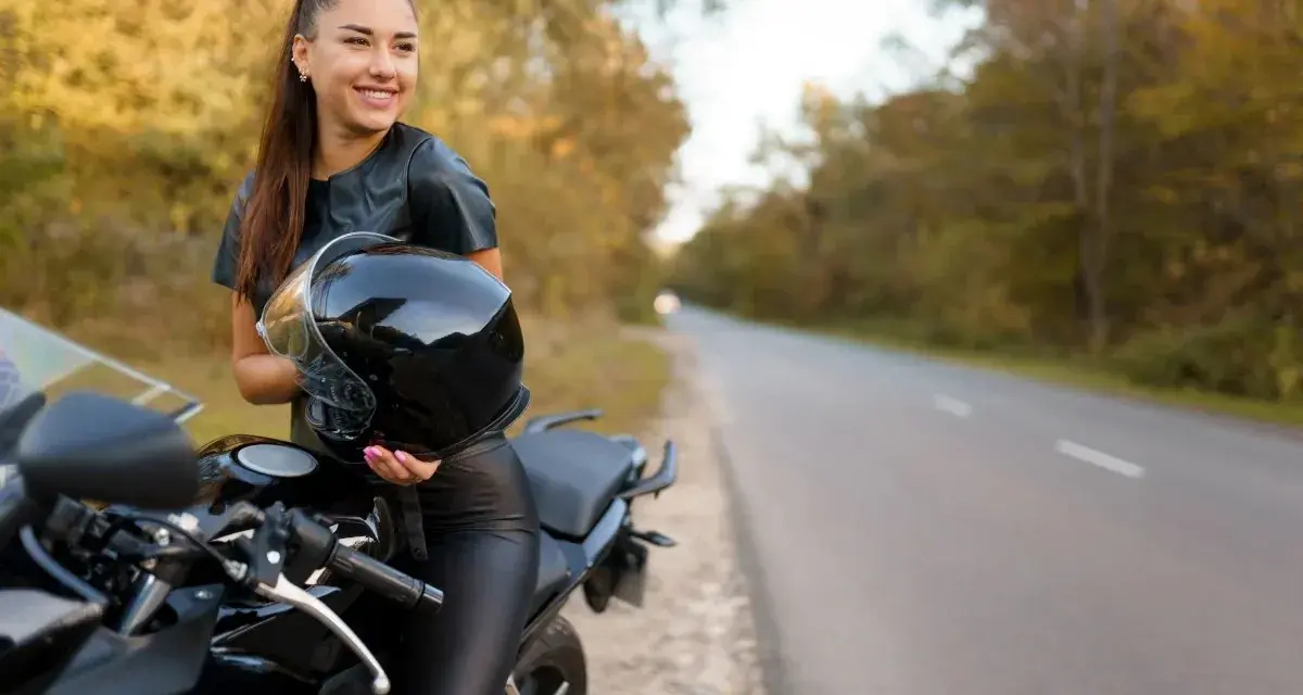 Uśmiechnięta kobieta w skórzanym stroju trzyma kask obok swojego motocykla. Idealne motory dla kobiet na jesienną przejażdżkę.