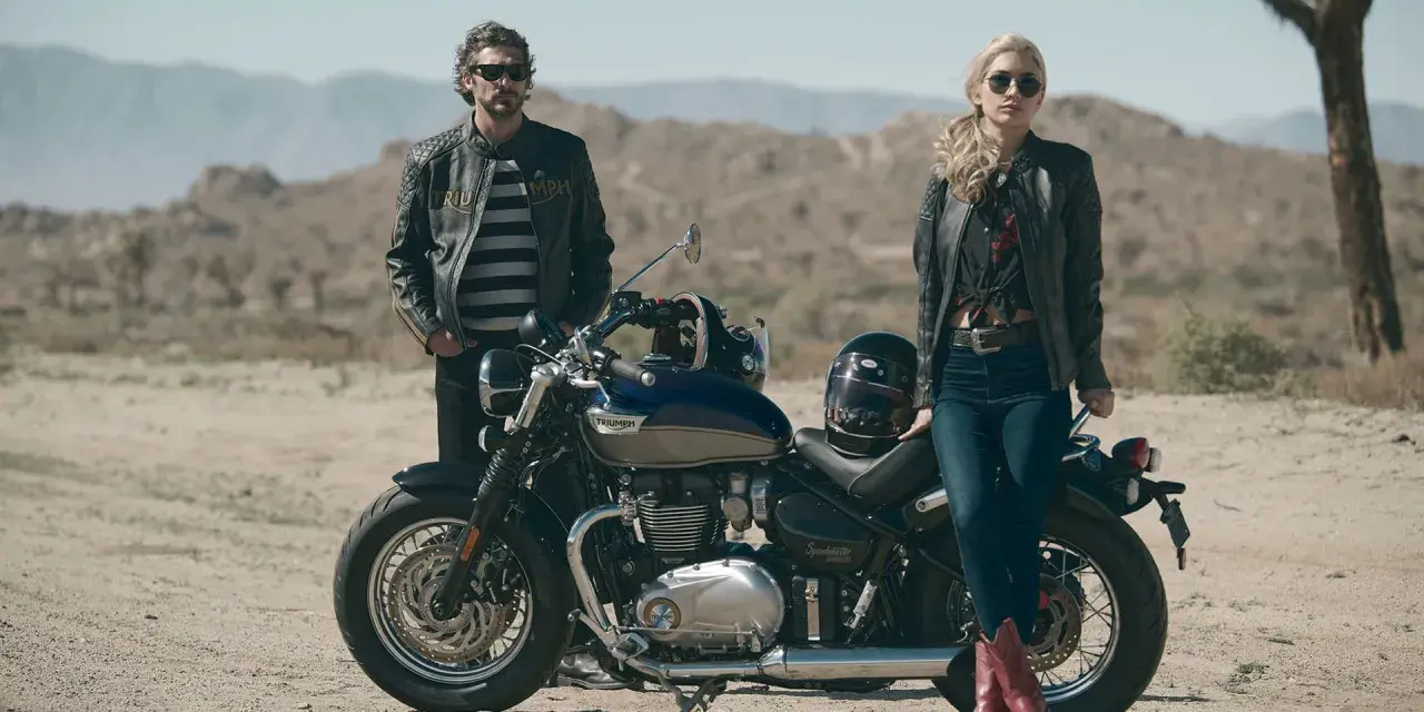 Para w skórzanych kurtkach pozuje przy motocyklu Triumph Speedmaster na pustynnym tle.
