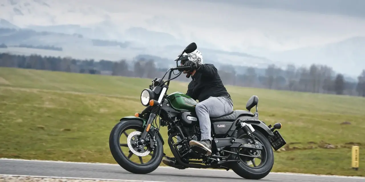 Motocyklista na zielonym chopperze UM Renegade 125 przemierza malowniczą drogę wśród zielonych pól i gór.