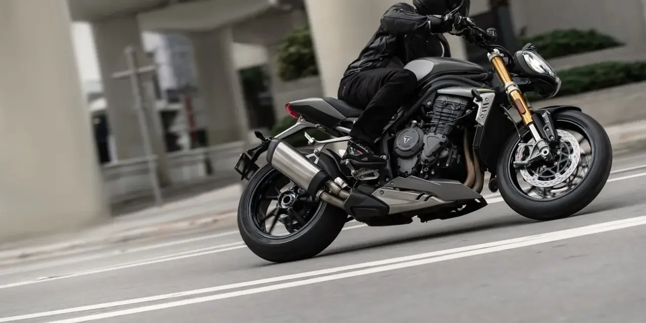 Motocyklista w kasku i skórzanej kurtce pochyla się na swoim motocyklu Triumph Speed Triple, pokonując zakręt na miejskiej drodze.