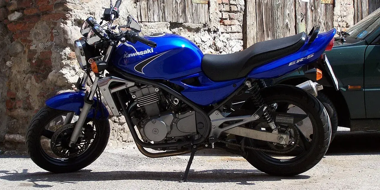 Błękitny motocykl Kawasaki ER-5 zaparkowany przy kamiennym murze i drewnianej bramie.