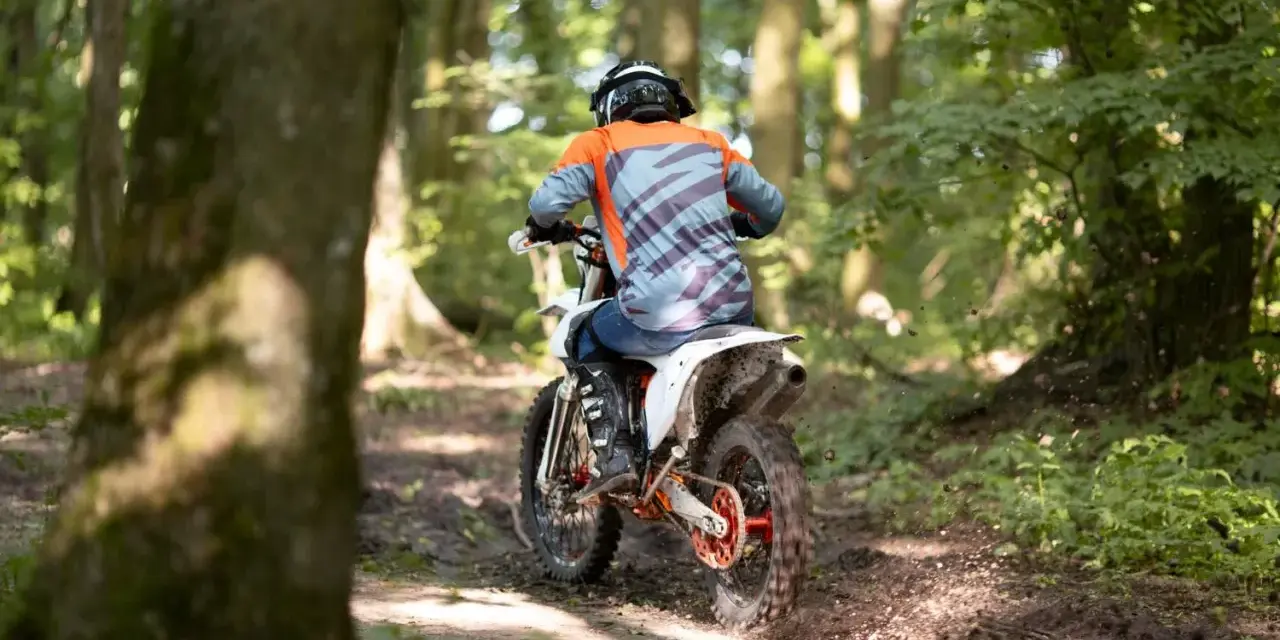 Motocyklista w kasku i kolorowej bluzie pędzi leśnym szlakiem. Od ilu lat można jeździć crossem? Odpowiedź zależy od przepisów i umiejętności.