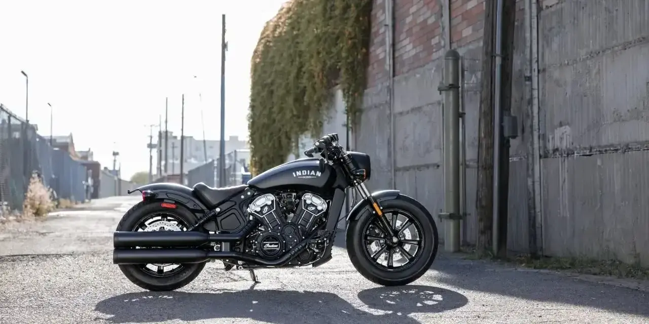 Czarny motocykl typu cruiser Indian Scout stoi na ulicy, z zielonym bluszczem zwisającym ze ściany w tle.