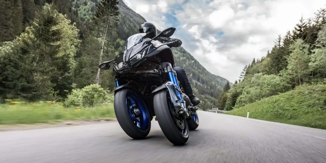 Motocyklista na Yamaha Niken pokonuje zakręt na górskiej drodze, otoczony zielonymi lasami i skalistymi szczytami.