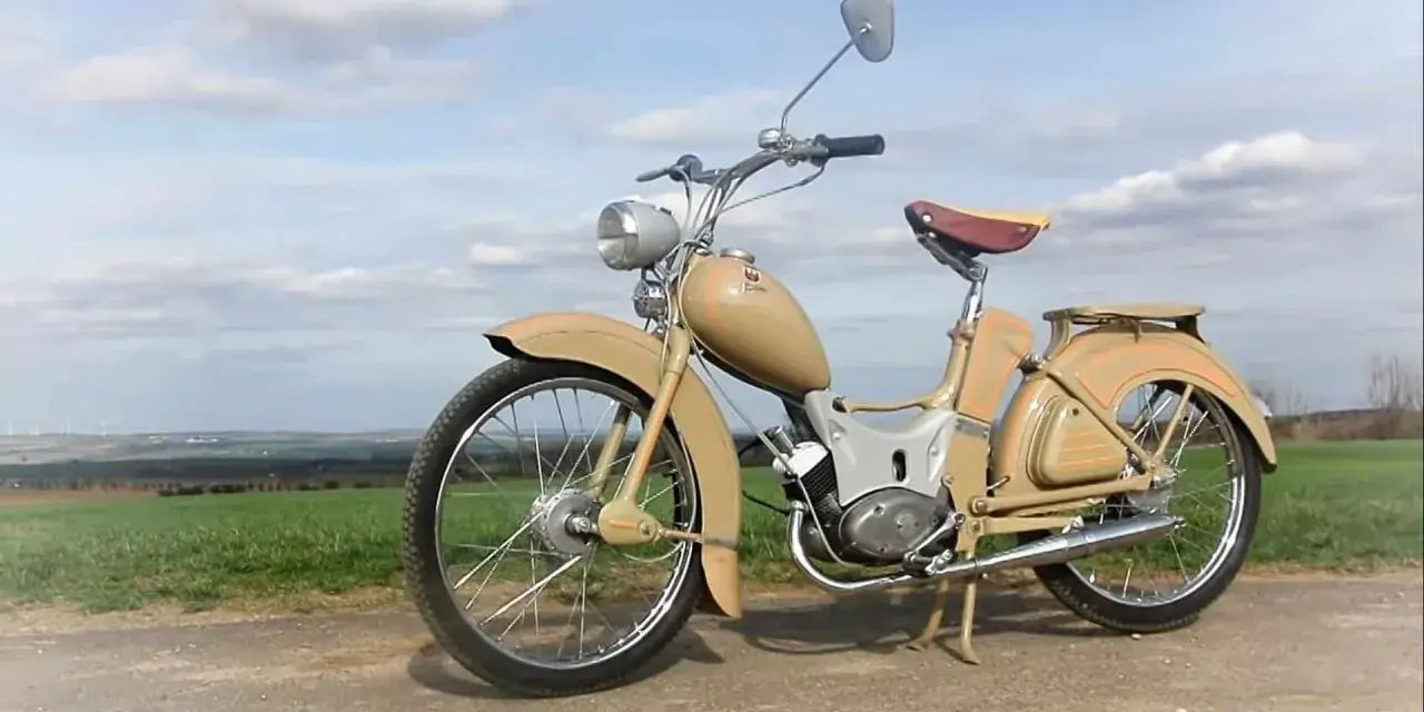 Beżowy motorower z NRD, z charakterystycznym siodełkiem i błotnikami, stoi na tle zielonych pól i błękitnego nieba.