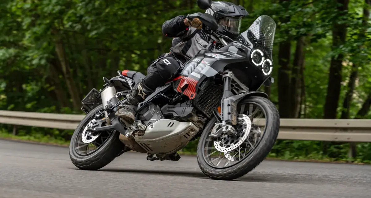 Motocyklista w kasku i stroju terenowym pochyla się na zakręcie na drodze leśnej, prowadząc czarny motocykl Ducati DesertX.