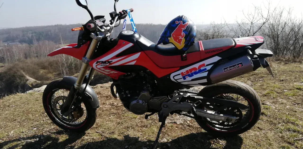 Czerwony motocykl Honda FMX 650 z kaskiem Red Bull na siedzeniu, gotowy na przygodę w terenie.