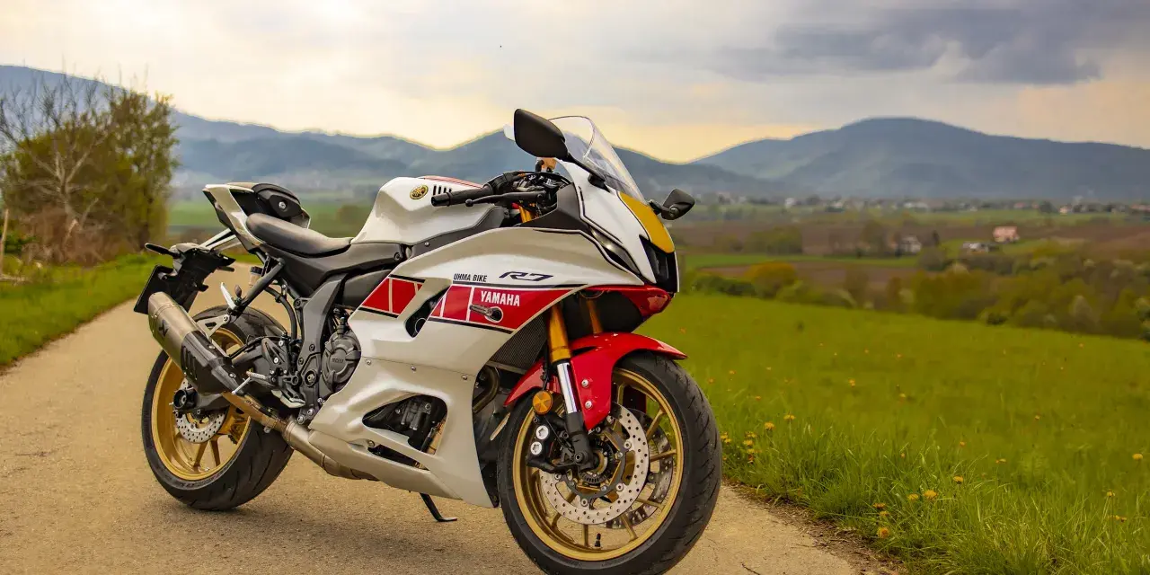 Sportowy motocykl Yamaha R7 w białym malowaniu z czerwonymi akcentami, zaparkowany na drodze wśród zielonych wzgórz. Sprawdź aktualną yamaha r7 cena!