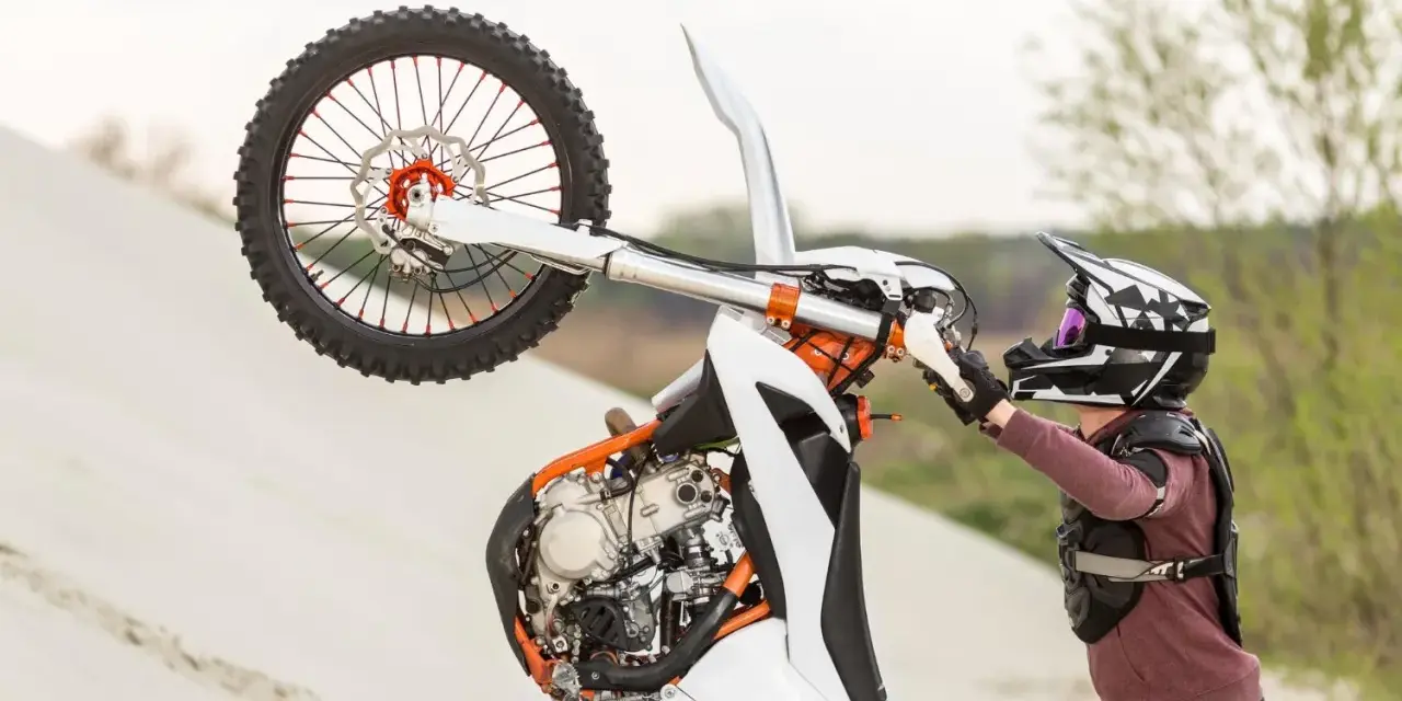 Motocyklista w kasku i ochraniaczach wykonuje wheelie na piaskowej wydmie. To najszybszy cross na świecie, gotowy do akcji.