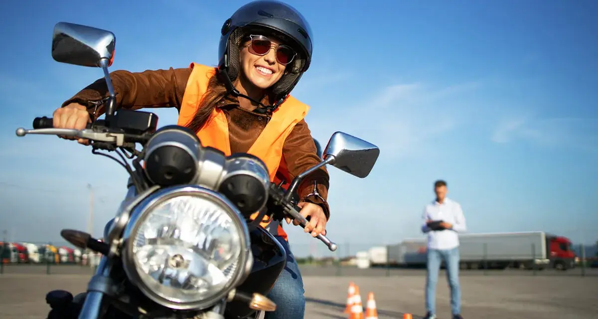Uśmiechnięta kursantka w kamizelce odblaskowej i kasku na motocyklu, przygotowuje się do egzaminu na prawo jazdy A2.