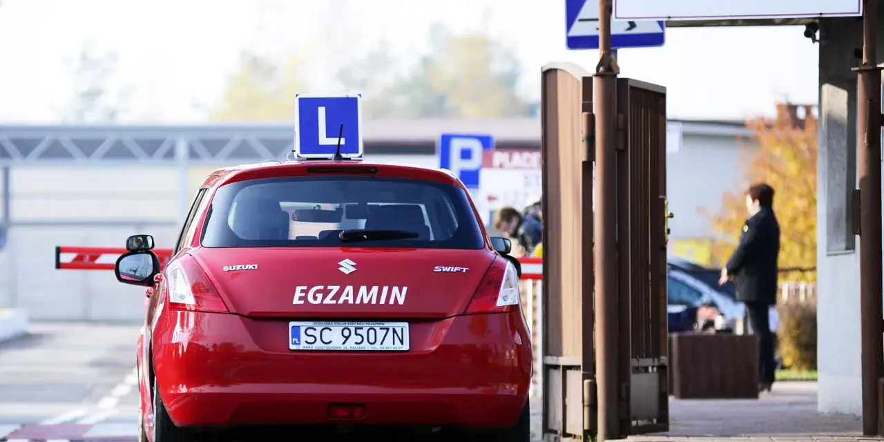 Czerwony samochód z literą "L" na dachu i napisem "EGZAMIN" czeka przed bramą. Czy trudno zdać prawo jazdy?