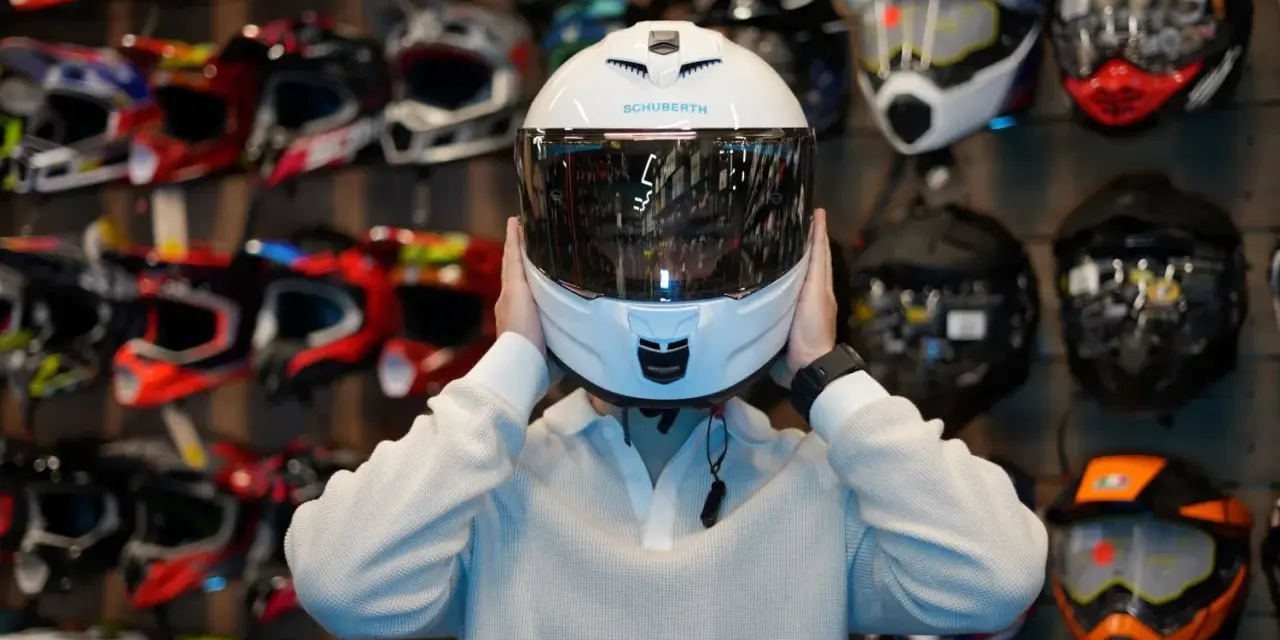 Osoba przymierza biały, cichy kask motocyklowy Schuberth. W tle widać ścianę pełną kolorowych kasków.