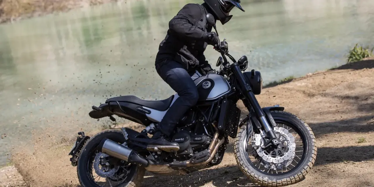 Motocyklista w kasku i czarnej kurtce jedzie motocyklem Benelli Leoncino 500 Trail po piaszczystym brzegu rzeki, wzbijając tumany kurzu.