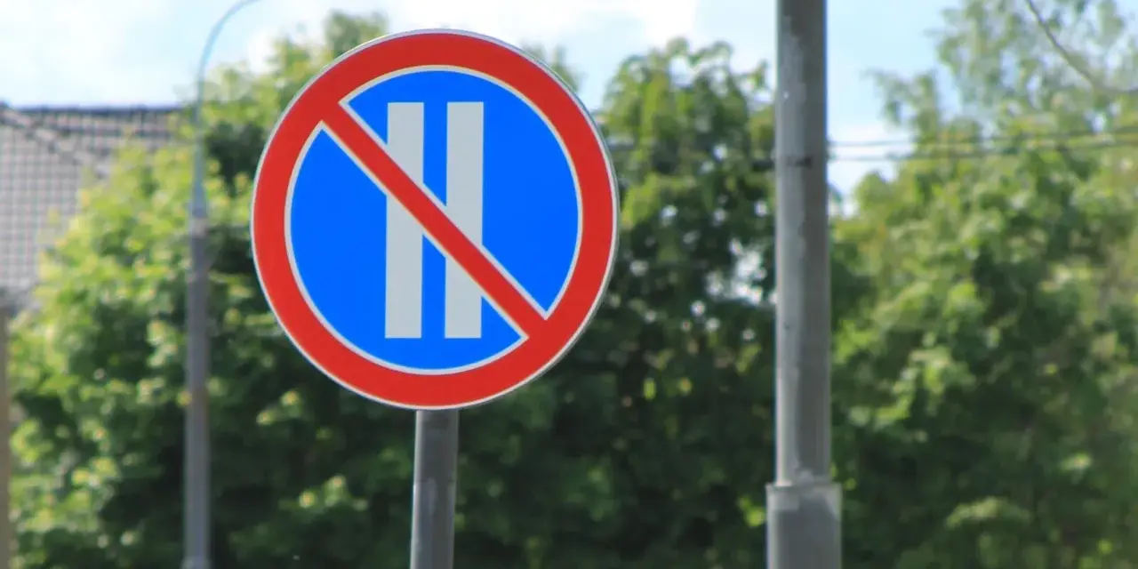 Znak B38 zakazujący postoju. Okrągły znak z czerwonym obramowaniem, niebieskim tłem i dwoma białymi pionowymi pasami przekreślonymi czerwoną linią.
