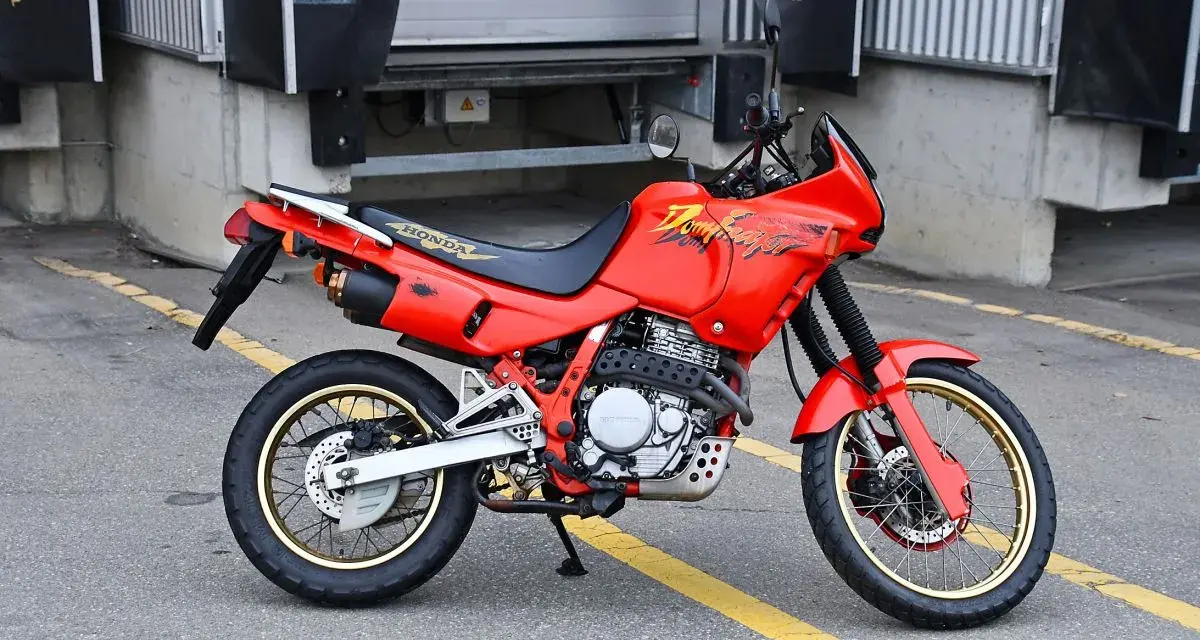 Czerwony motocykl Honda Dominator z lat 90. stoi na asfalcie. Ma złote felgi i czarne siedzenie.